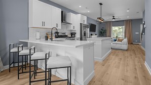 A modern kitchen with white cabinetry, a central island with bar stools, a built-in oven, and a large window in the adjacent living area.