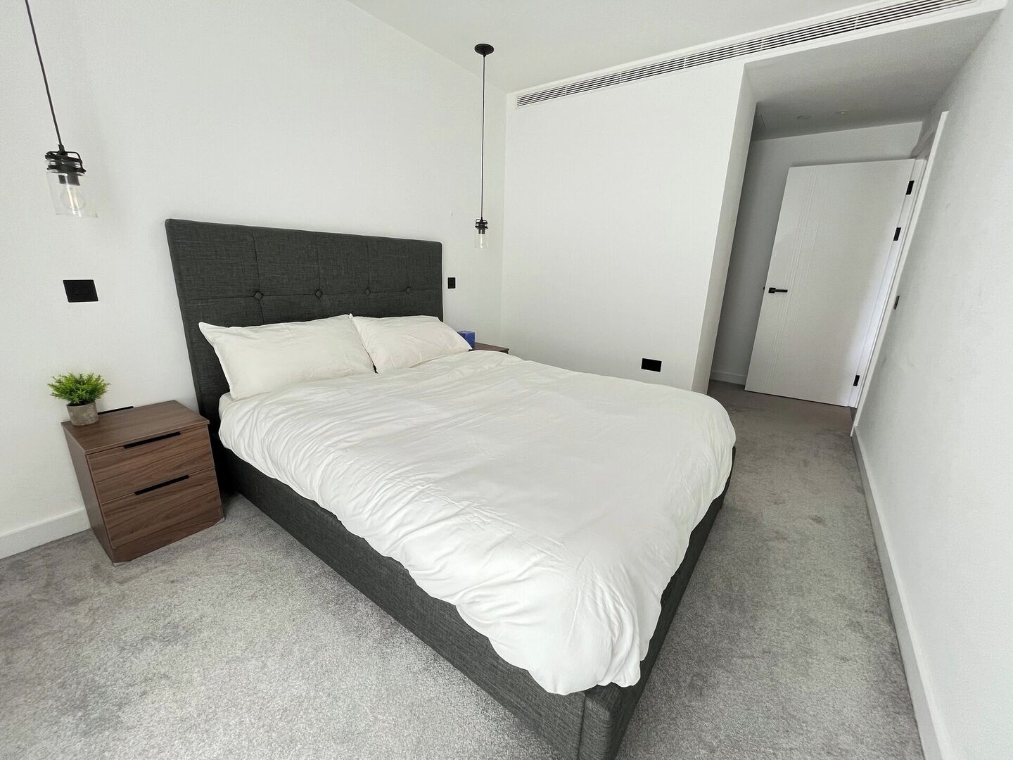 A modern bedroom with a large bed, a nightstand, a potted plant, and a door leading to another room.