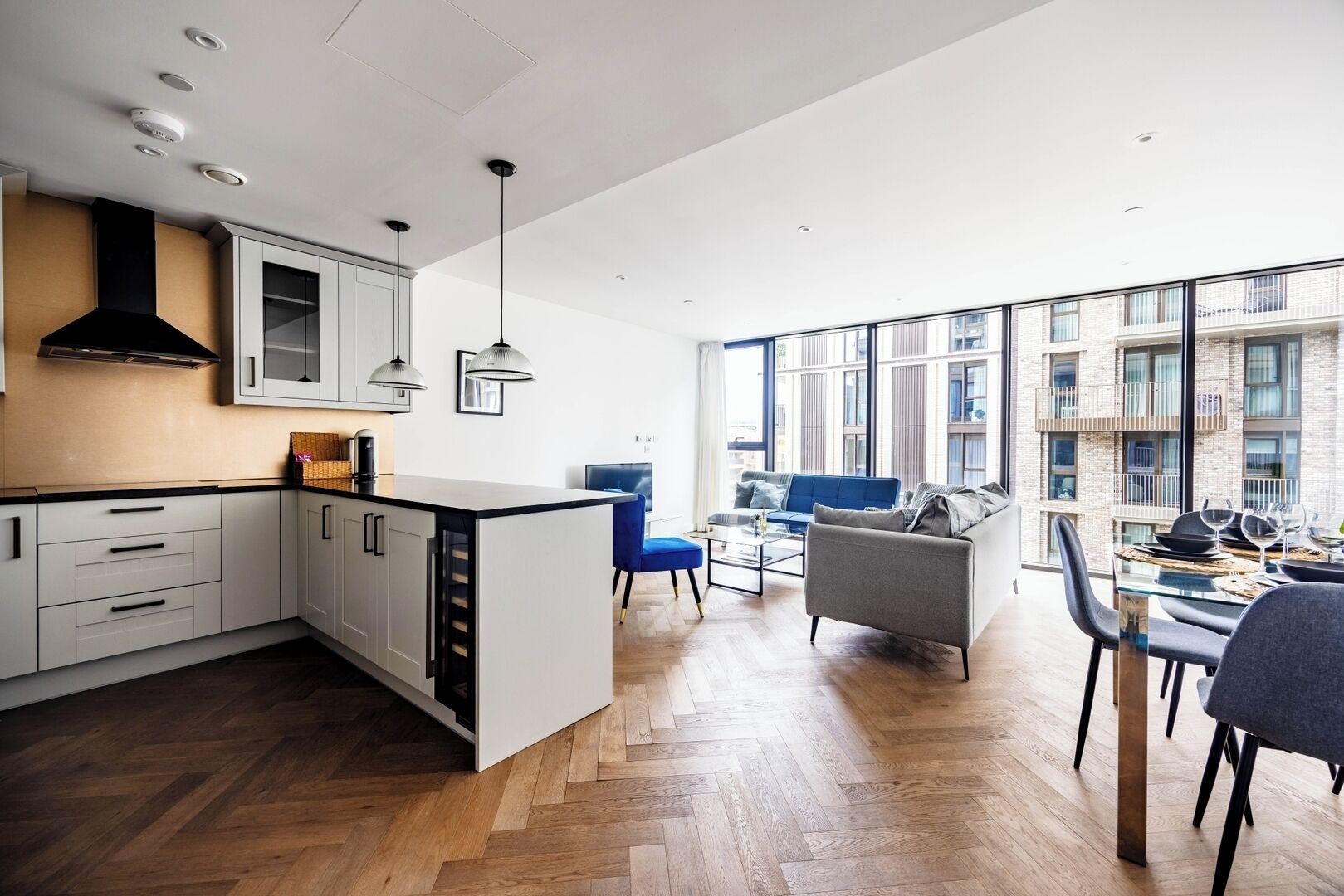 Une cuisine moderne avec des armoires blanches, une hotte noire et des luminaires suspendus. On y trouve un coin repas avec une table et des chaises en verre, ainsi qu’un canapé. De grandes fenêtres offrent une vue sur l’extérieur du bâtiment.