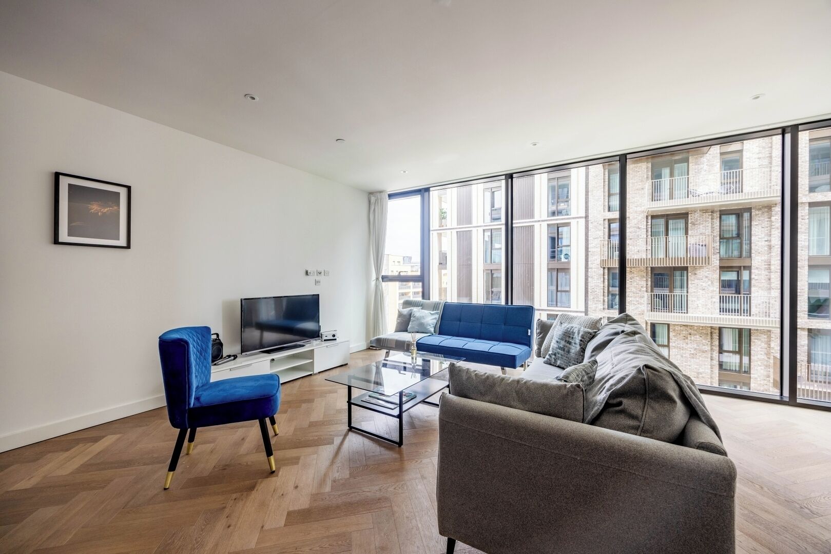 Un salon moderne avec une grande fenêtre, un téléviseur à écran plat, un fauteuil bleu, un canapé gris et une table basse en verre.