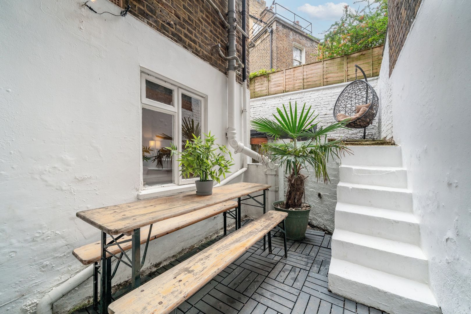 A narrow outdoor patio with a wooden bench, a potted plant, and a hanging chair.