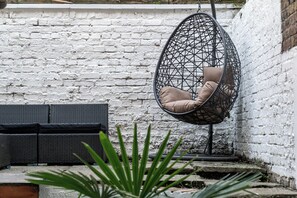 Une chaise suspendue avec un coussin, adossée à un mur de briques blanches, un canapé noir en osier et des plantes vertes au premier plan.