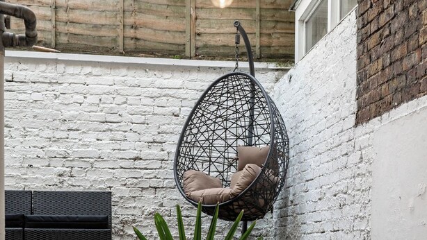 A hanging chair with cushions, a potted plant, and a staircase against a white brick wall. - Cozy Nook • Wood Burner • Terrace (London)