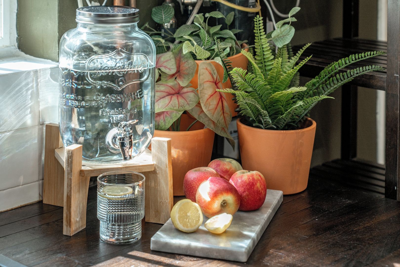 A clear glass water dispenser, a potted plant, apples, and a glass on a wooden stand.