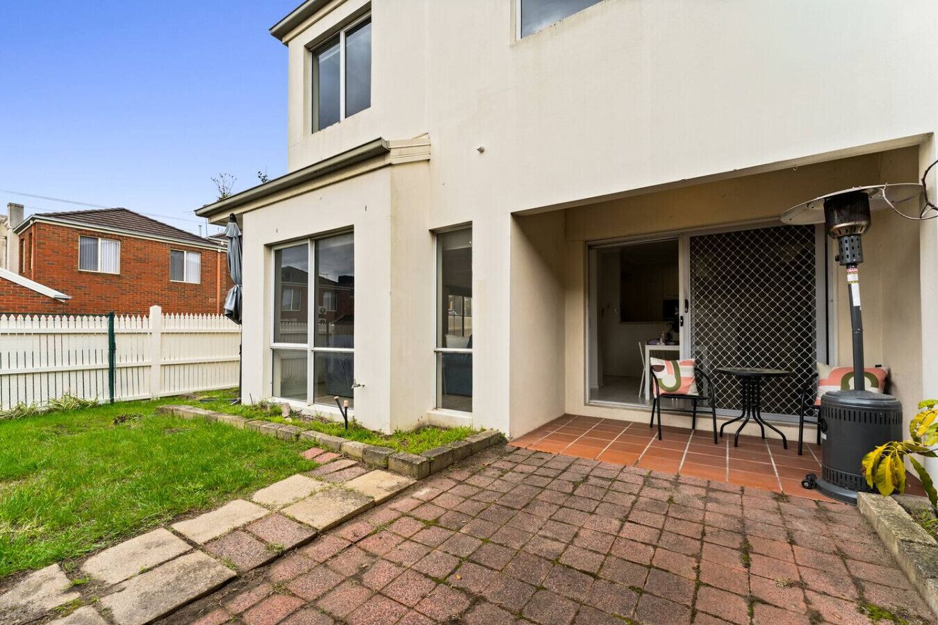 A two-story house with a patio area featuring a table and chairs, a sliding glass door, and a white picket fence.