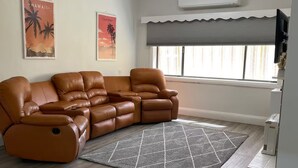 Una sala de estar con un sofá de cuero marrón, un ventilador de techo, una ventana con persianas y dos carteles en la pared.