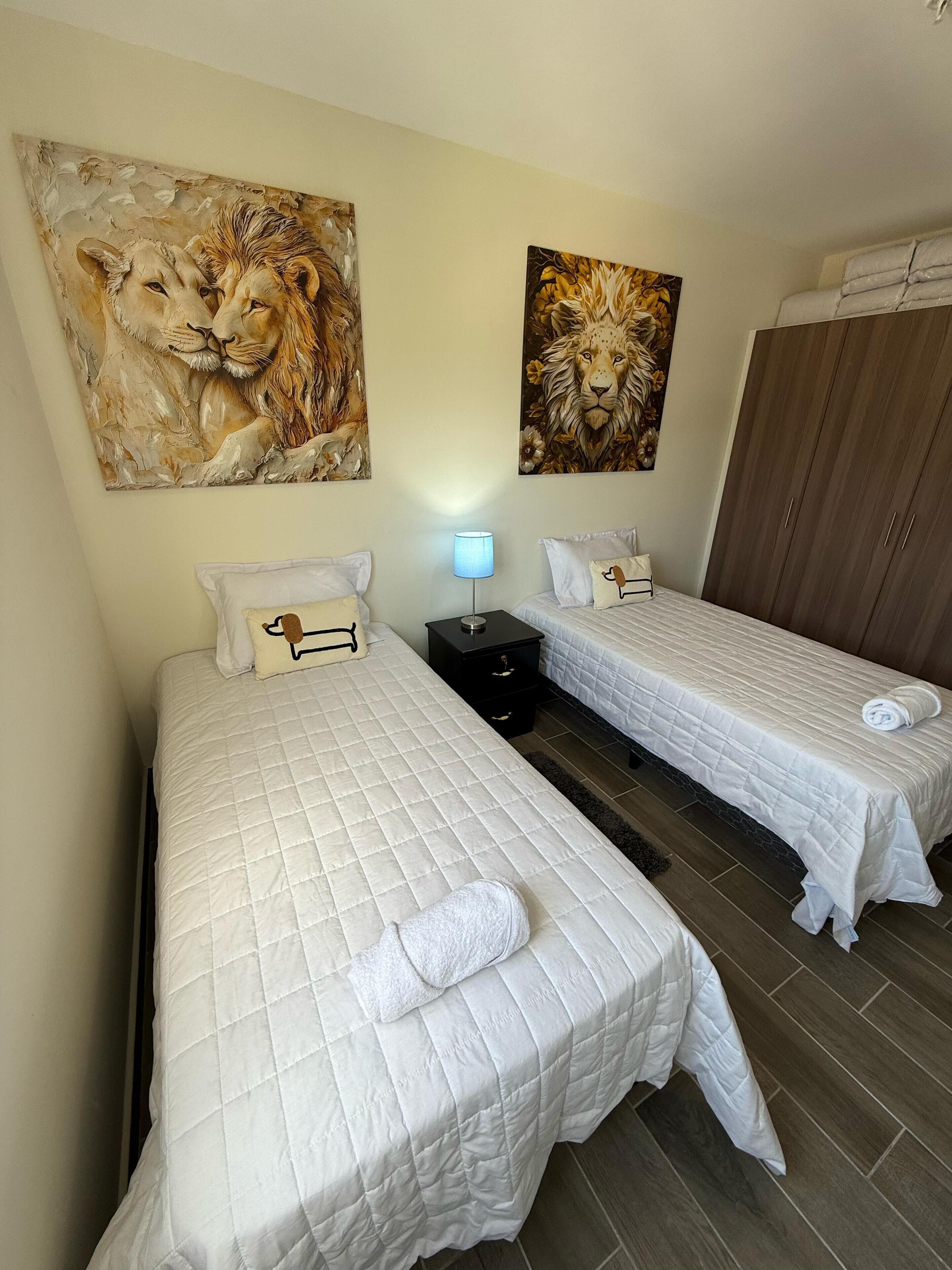 Habitación de hotel con dos camas, una mesita de noche con lámpara y dos cuadros de leones en la pared.