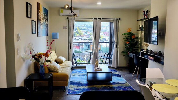 Een moderne woonkamer met een bank, salontafel en een eethoek met stoelen. Er is een balkon met uitzicht op gebouwen.