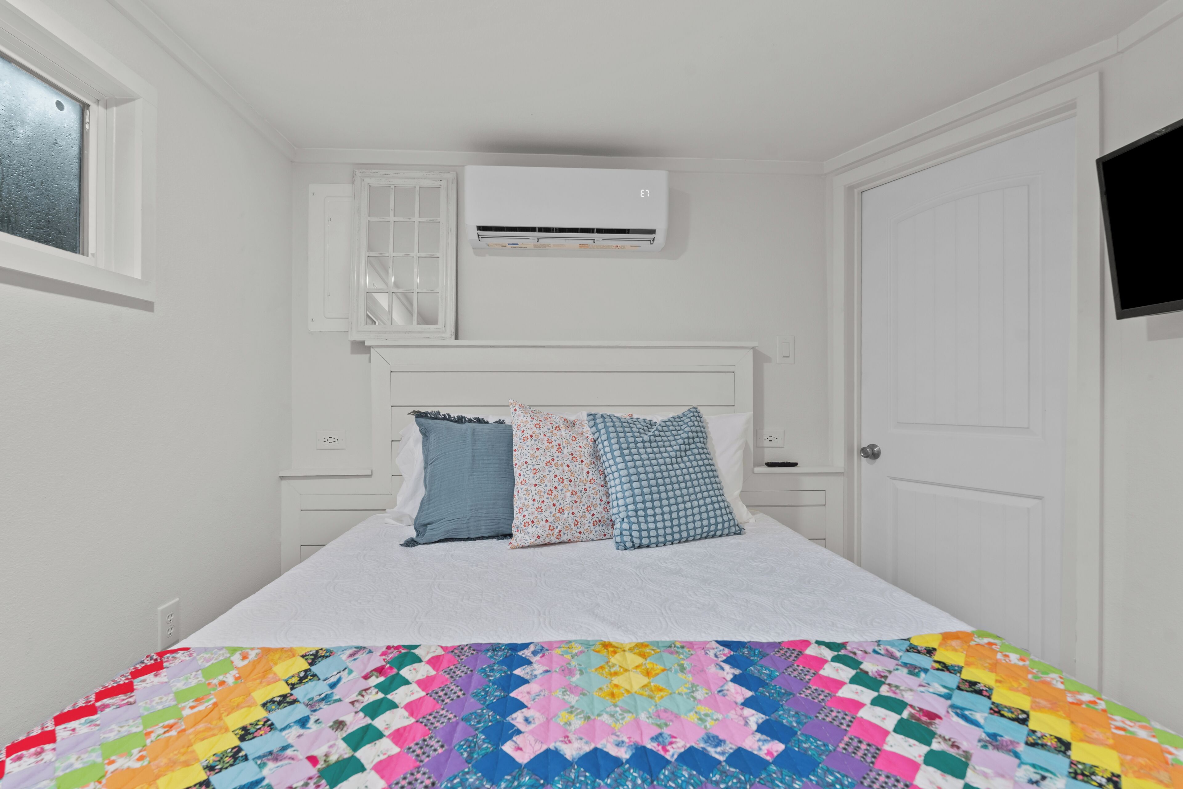 Ein Schlafzimmer mit einem Bett, weißen Wänden, einem Fenster, einer Klimaanlage und einer Tür.