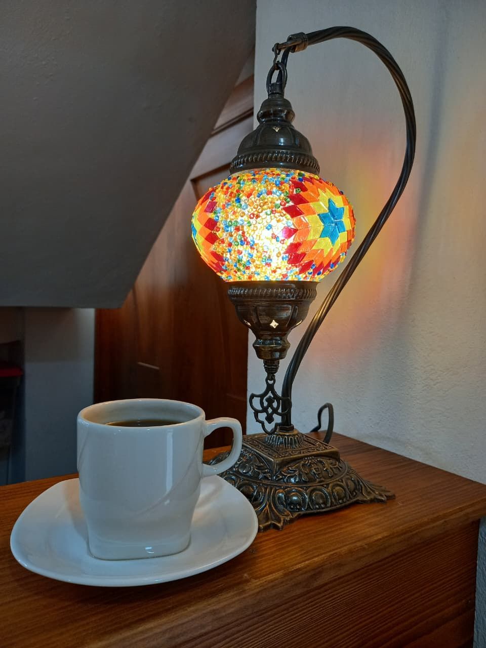 A white cup of coffee on a saucer next to a decorative lamp with a metal base and a colorful glass shade.