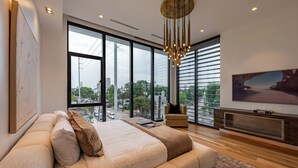 A modern bedroom with a large bed, a seating area, a television, and a city view. - The Glass Haus II· Modern Luxury · River Oaks · Rooftop Terrace · Elevator ⭐️⭐️⭐️⭐️⭐️ (Houston)