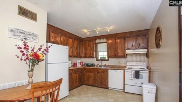 A compact kitchen with wooden cabinets, a white refrigerator, a stove, and a sink. There is a dining table with chairs and a vase of flowers on the wall. - Cozy 2-bedroom townhome in awesome Columbia with AC. (Columbia)
