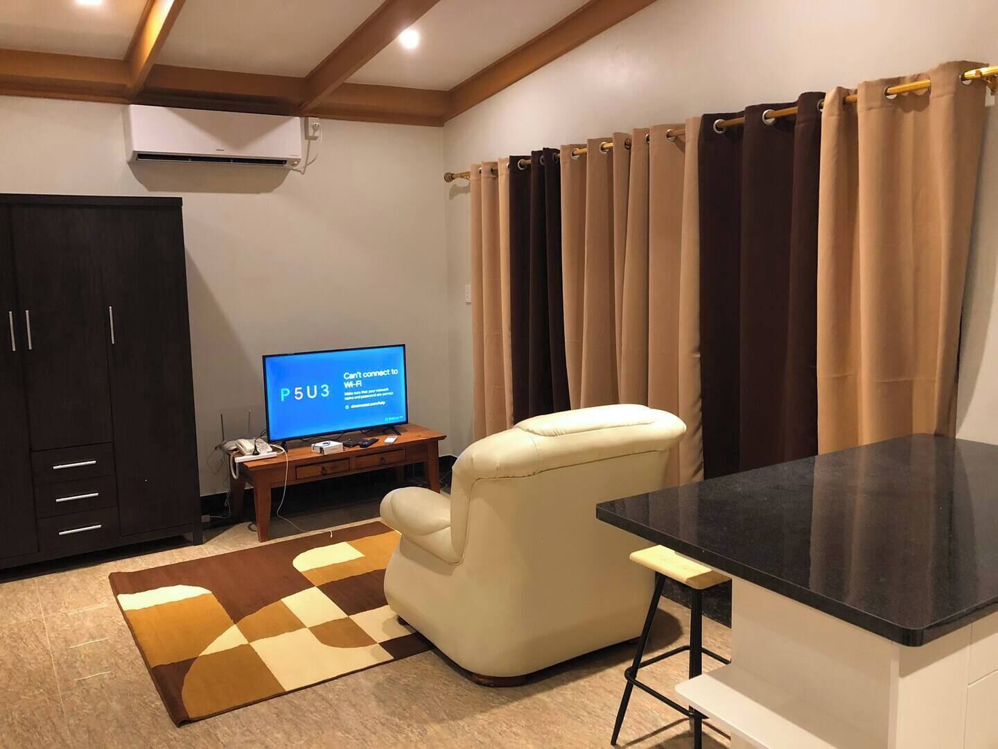 Un salon avec un téléviseur à écran plat affichant un message d’erreur concernant la connexion Wi-Fi, un fauteuil beige, une petite table d’appoint en bois et un tapis à carreaux.