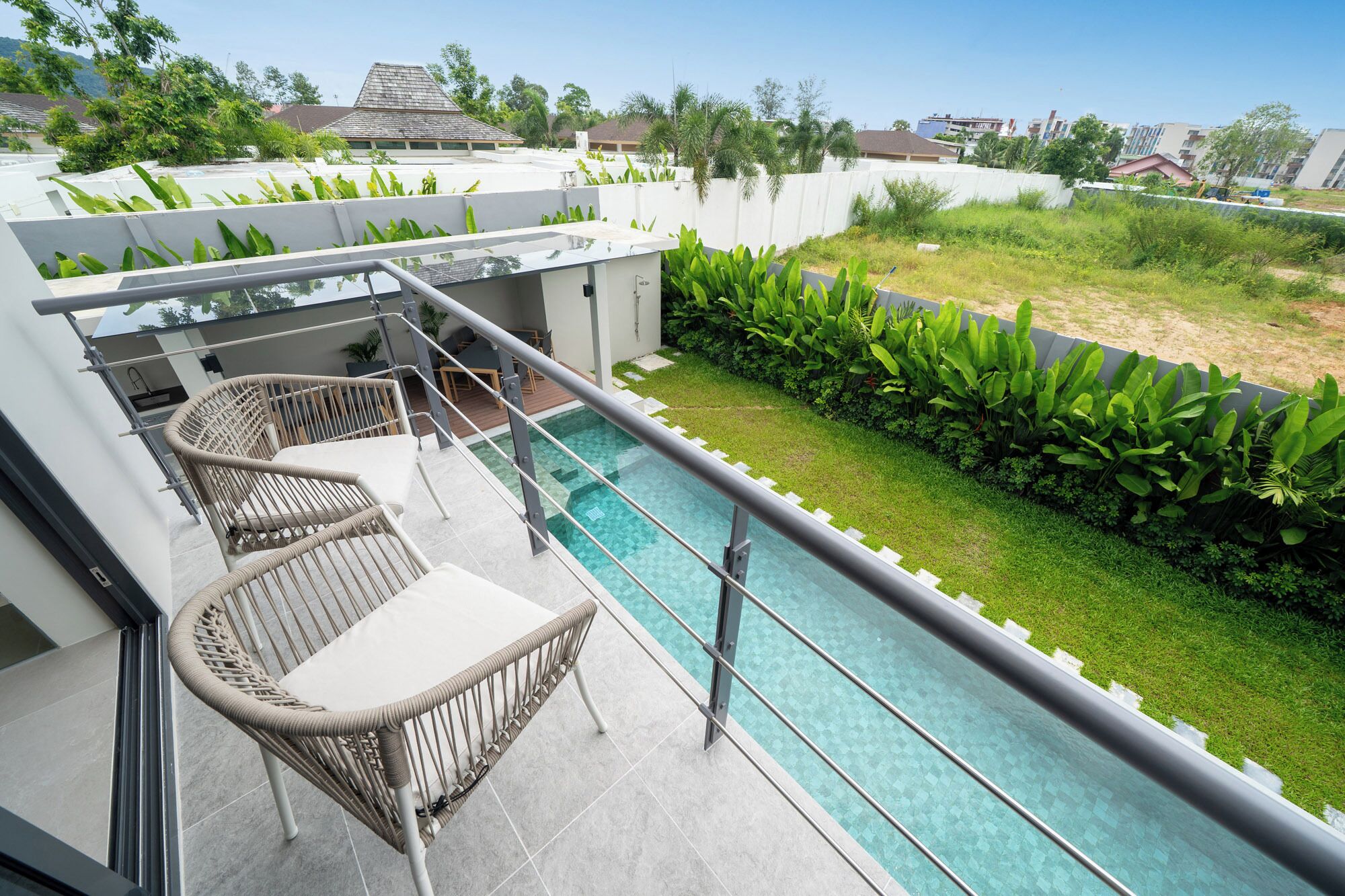 Un área de piscina en la azotea con barandilla de vidrio, dos sillones de mimbre y vistas a la vegetación y edificios circundantes.