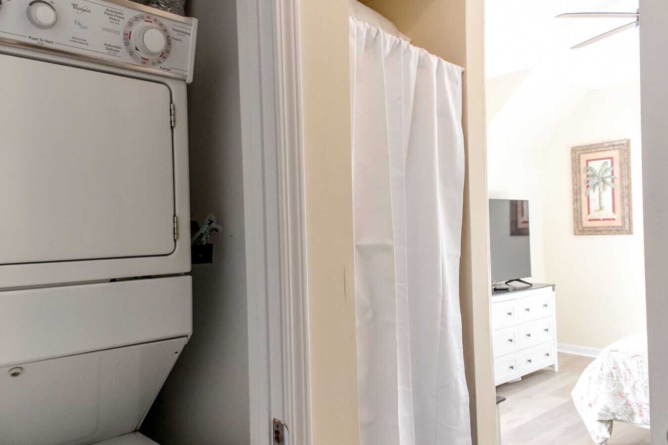 Uma lavanderia com máquinas de lavar e secar empilhadas, uma cortina branca e um quarto com cômoda e um quadro na parede.