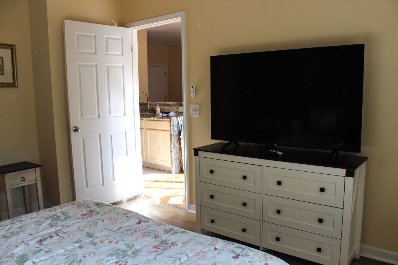 A bedroom with a bed, a dresser, a television, and an open door leading to a kitchen.