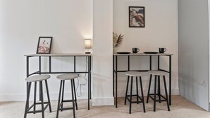 A small, modern kitchen with two bar stools, a framed picture, and a wall-mounted light.