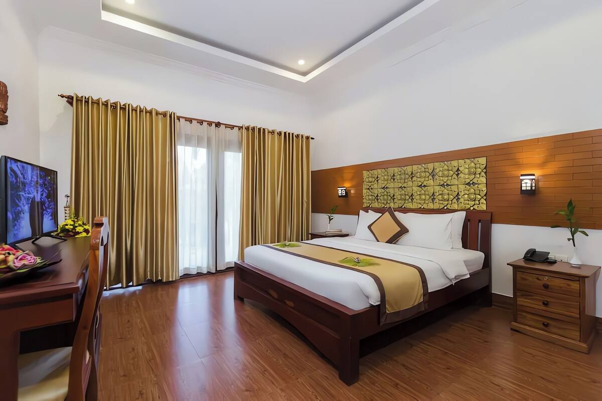 A hotel room with a large bed, wooden floor, curtains, a desk with a computer, and a nightstand with a phone.