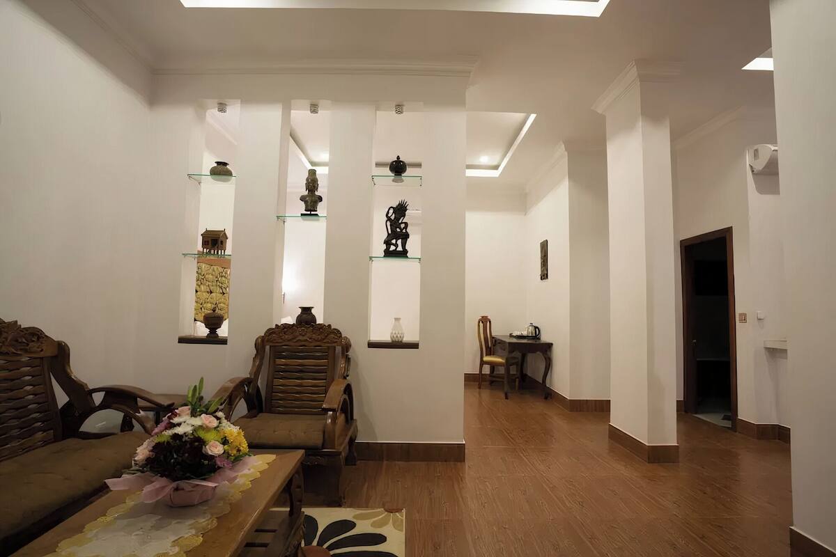 A spacious living room with a wooden dining table, two chairs, a sofa, and a floral arrangement. There are shelves with decorative items and a door leading to another room.