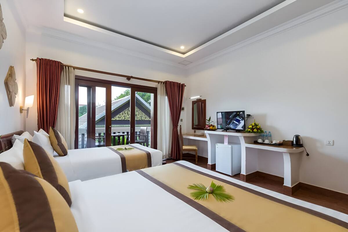 A hotel room with two beds, a desk with a TV, and a balcony with a view of a building.