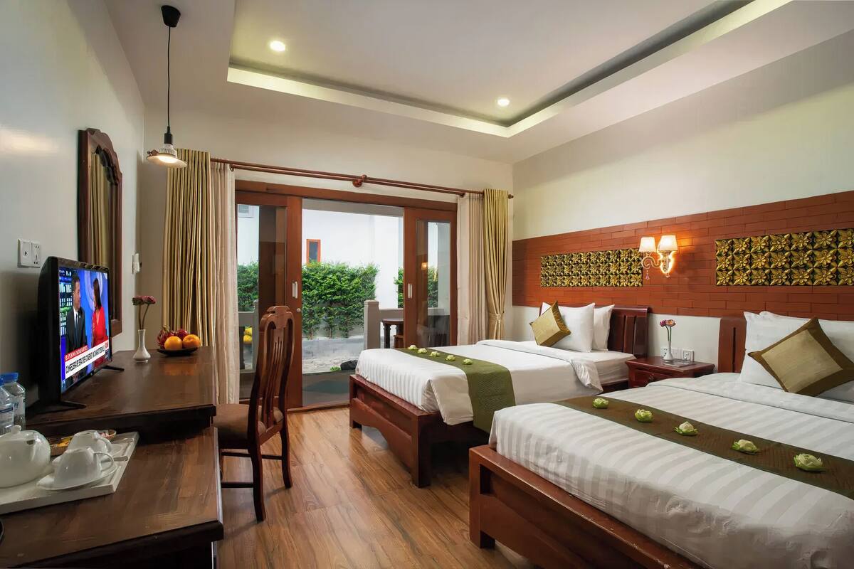 A hotel room with two beds, a desk with a TV, and a view of greenery outside.