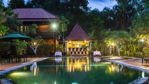 A pool area with a bar and seating, surrounded by lush greenery and a building with a red roof. - De Arya Angkor Resort (Siem Reap)