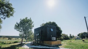 Une cabane moderne en bois, à deux étages, dotée d’une petite terrasse et entourée d’un champ verdoyant et d’arbres.