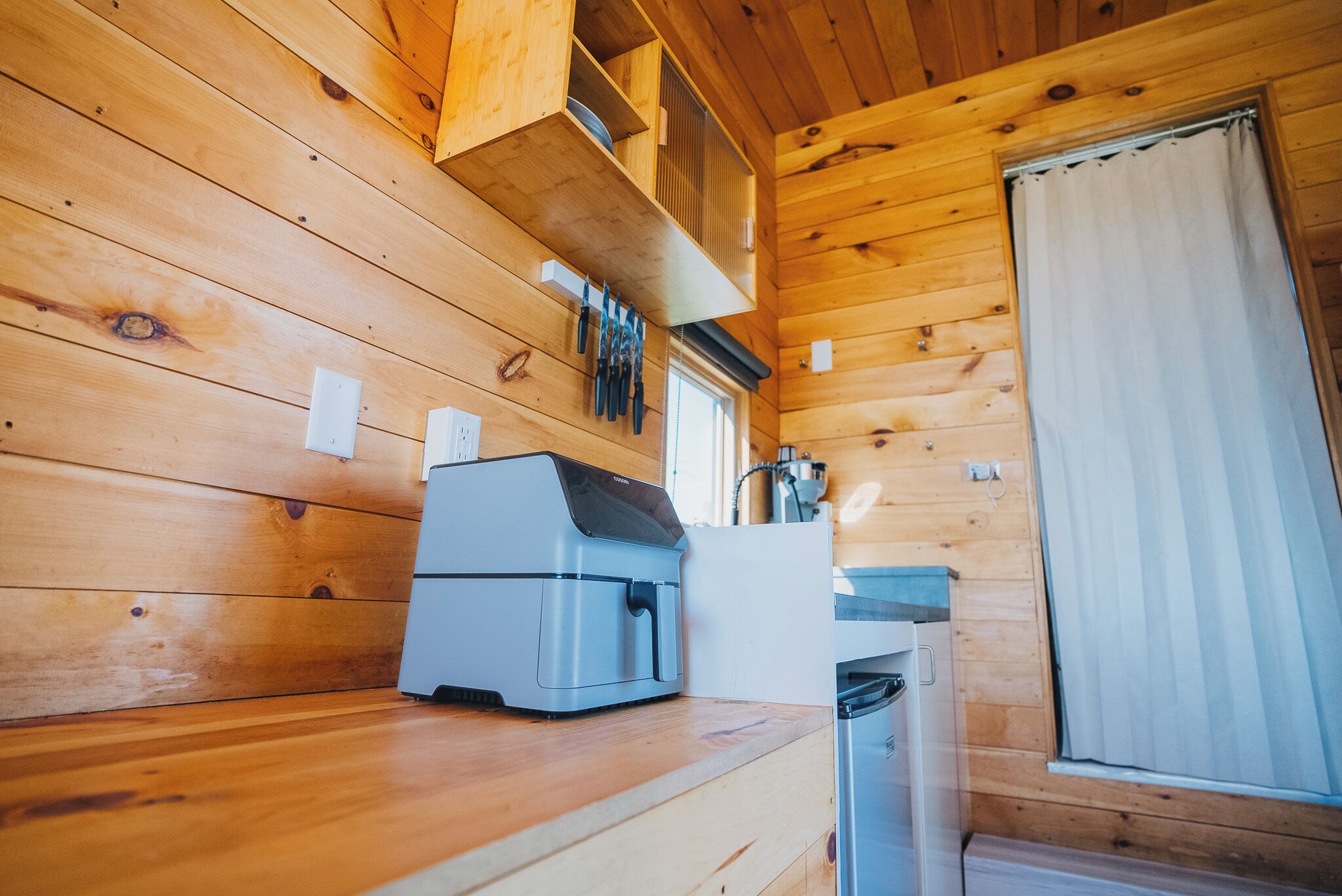 Interior de una cabaña de madera con cafetera, un lavabo y una ventana con cortinas.