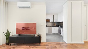 A modern living room with a flat-screen TV, a potted plant, and a wooden floor. A kitchen is visible in the background with white cabinets and a microwave. - GuestReady - Lovely Urban Retreat (Porto)