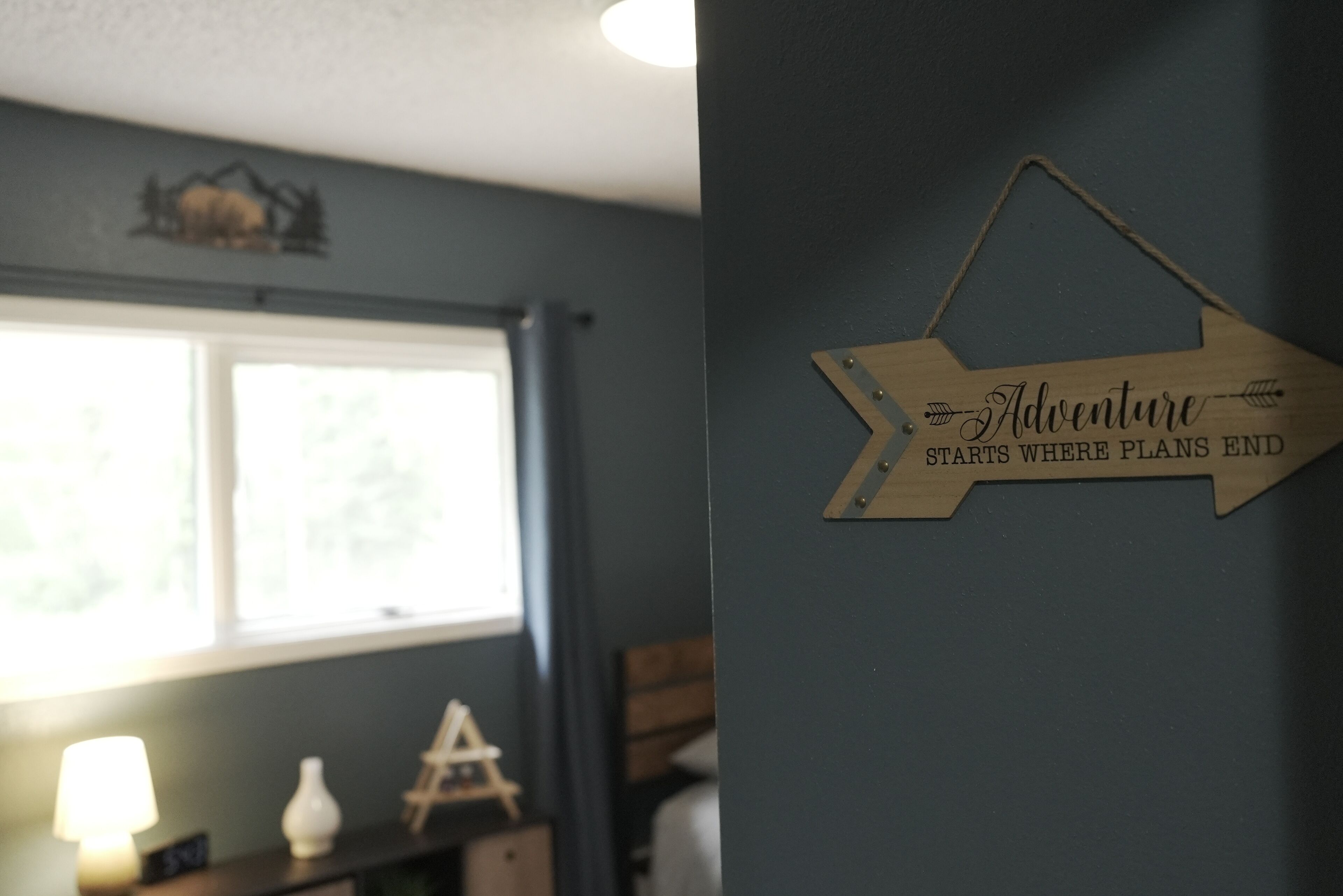 A room with a window, a lamp, and a wooden sign on the wall.