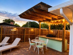 A patio with a wooden fence, a table with chairs, a barbecue grill, and a covered area with string lights.