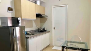 A compact kitchen with a stainless steel refrigerator, a microwave, and a stove. There is a glass table with a metal frame and a white door with a vent.