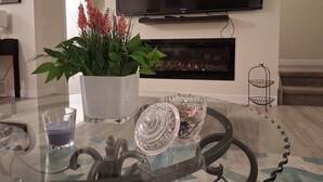 Un salon avec une table basse en verre, une plante en pot, un porte-bougie en verre et un bol décoratif en verre. Un téléviseur à écran plat est fixé au-dessus d’une cheminée.