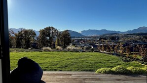 Une vue depuis une fenêtre donnant sur un espace verdoyant avec une terrasse en bois, des arbres et, au loin, une ville dont les montagnes se dessinent à l'horizon.