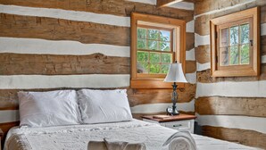 A log cabin bedroom with a wooden bed, white bedding, a nightstand with a lamp, and a window with a view of trees.