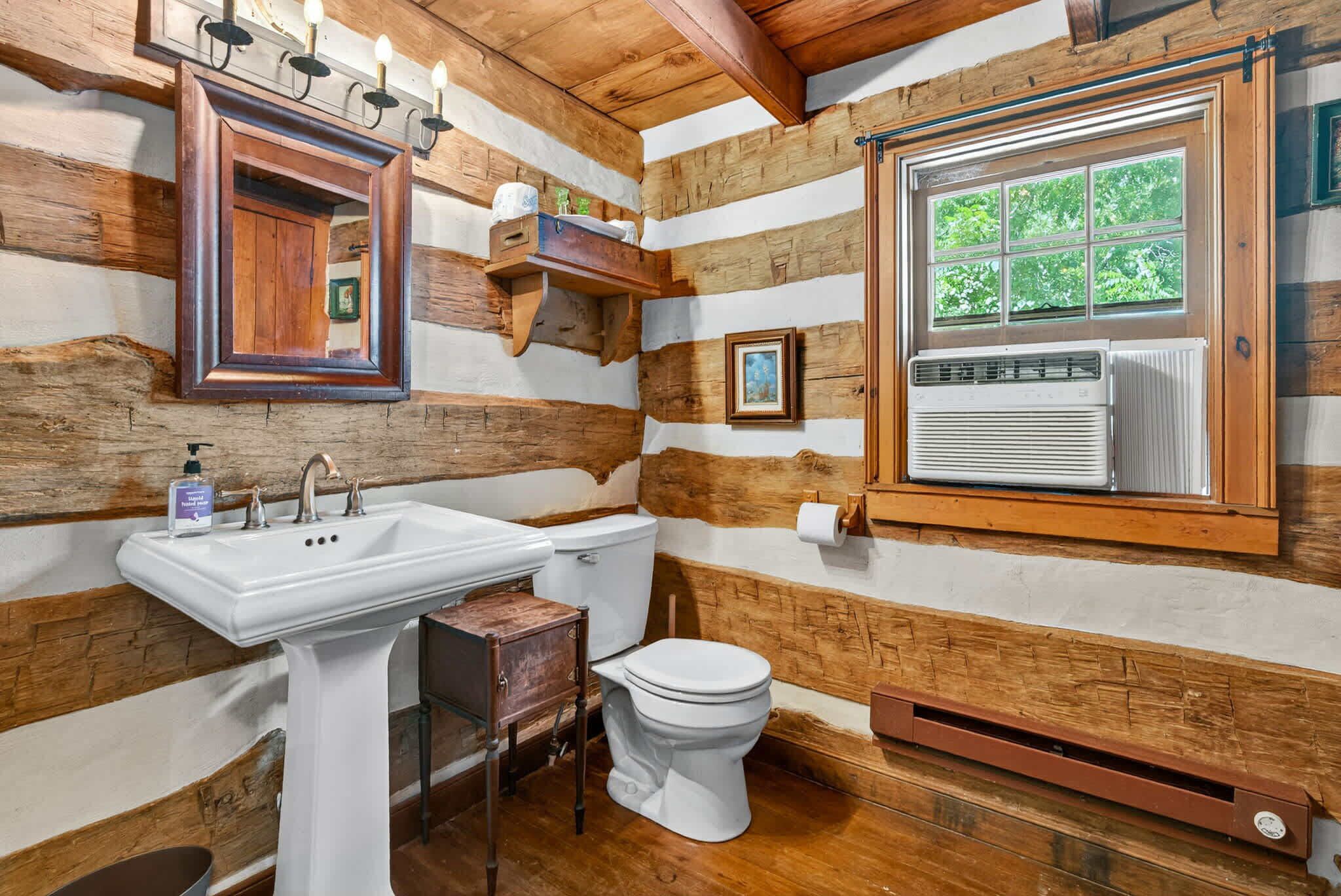 Un baño rústico con pared de madera, un lavamanos blanco, un inodoro y una ventana con vistas al verdor.