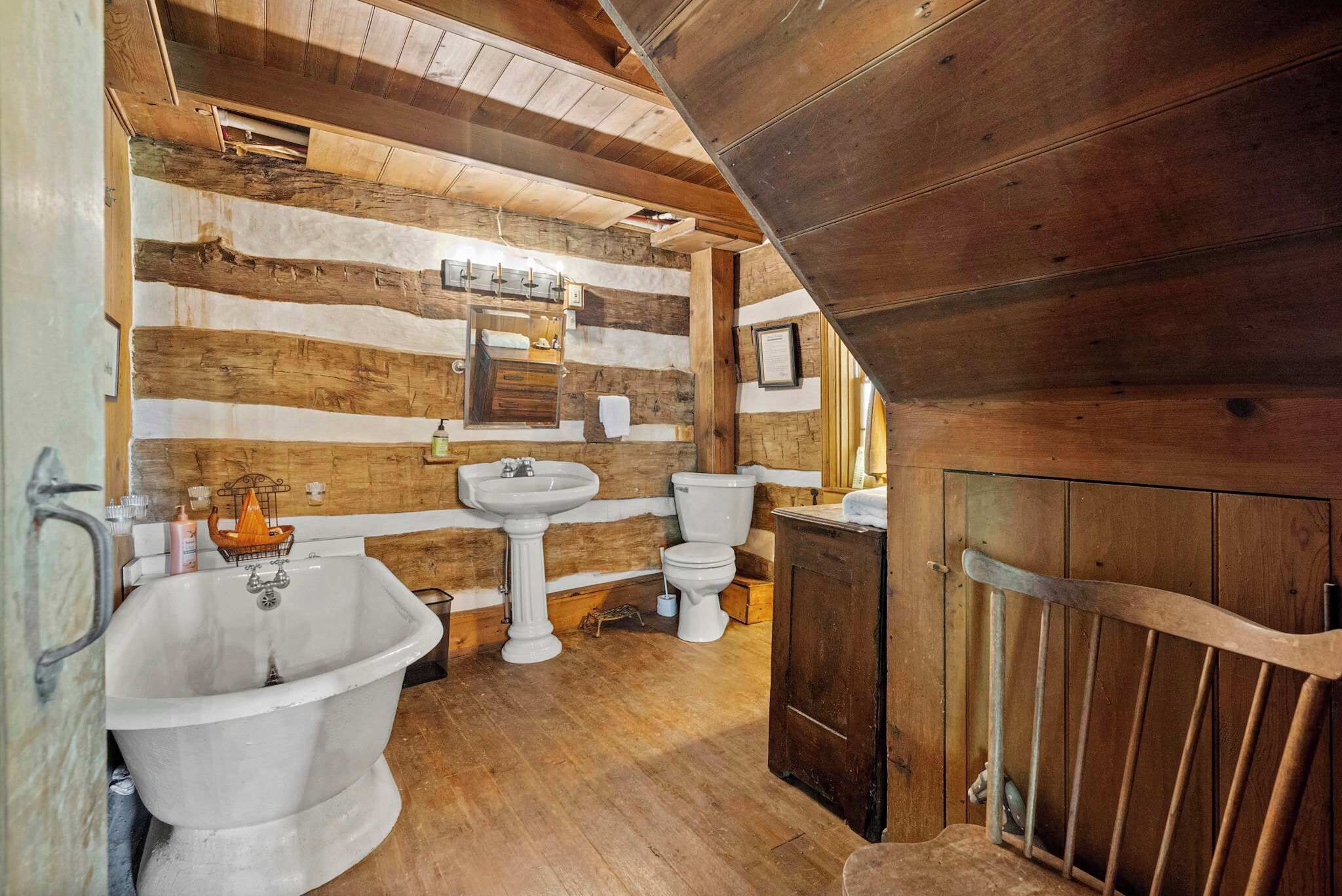 Un baño rústico con paredes de madera, una bañera tipo patas de león, un lavabo sobre pedestal y un inodoro.