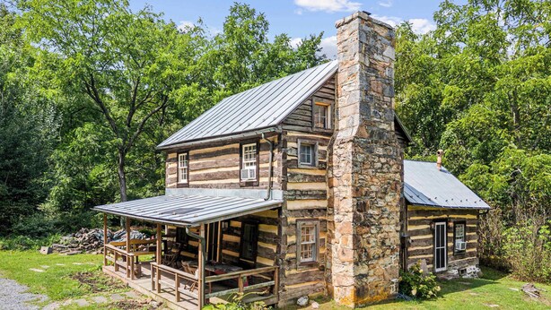 A log cabin with a stone chimney, a porch, and a small attached shed.