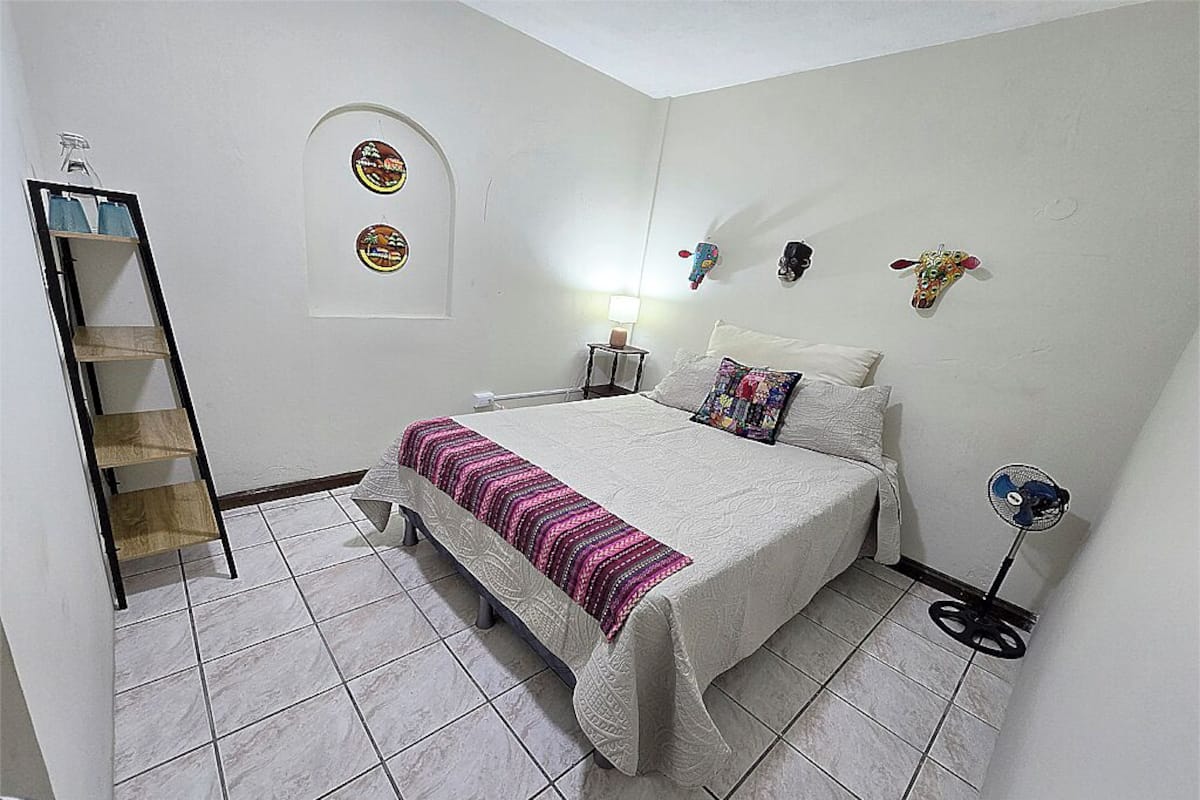 Une chambre à coucher comprenant un lit, une table de chevet, une lampe, un ventilateur fixé au mur et une étagère.
