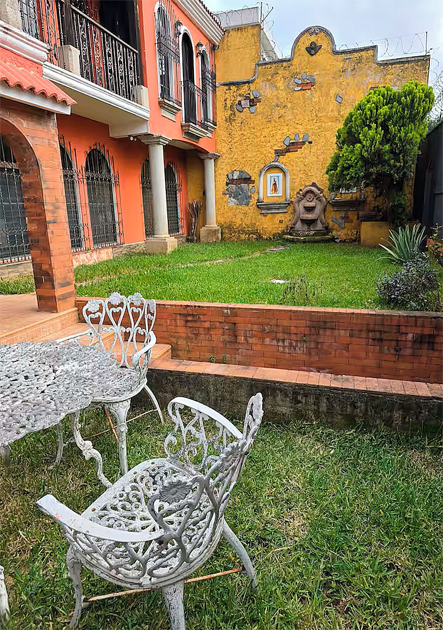 Un patio con una mesa y sillas de metal, un muro de ladrillo y un edificio amarillo con balcones y detalles decorativos.