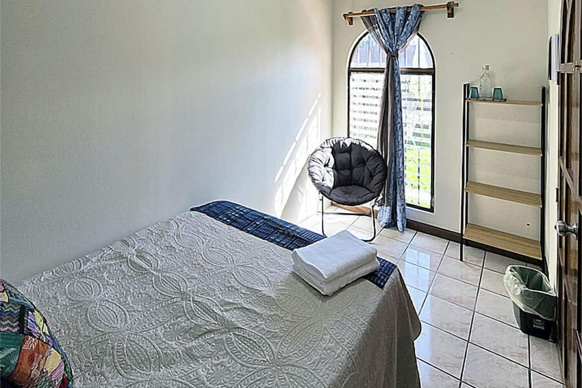 Une chambre à coucher avec un lit, une chaise, une fenêtre avec des rideaux, une étagère et un luminaire au plafond.