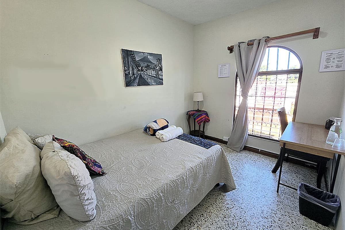 Une chambre à coucher comprenant un lit, un bureau, une chaise, une fenêtre avec des rideaux, une lampe fixée au mur et un tableau accroché au mur.