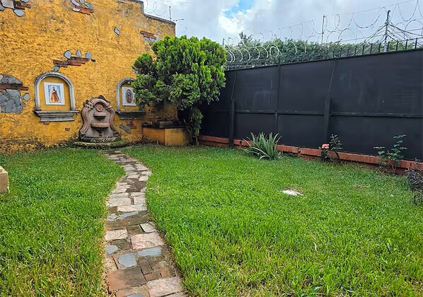 Un sendero de piedra que lleva a un patio con un muro amarillo, una cerca negra y un pequeño árbol.