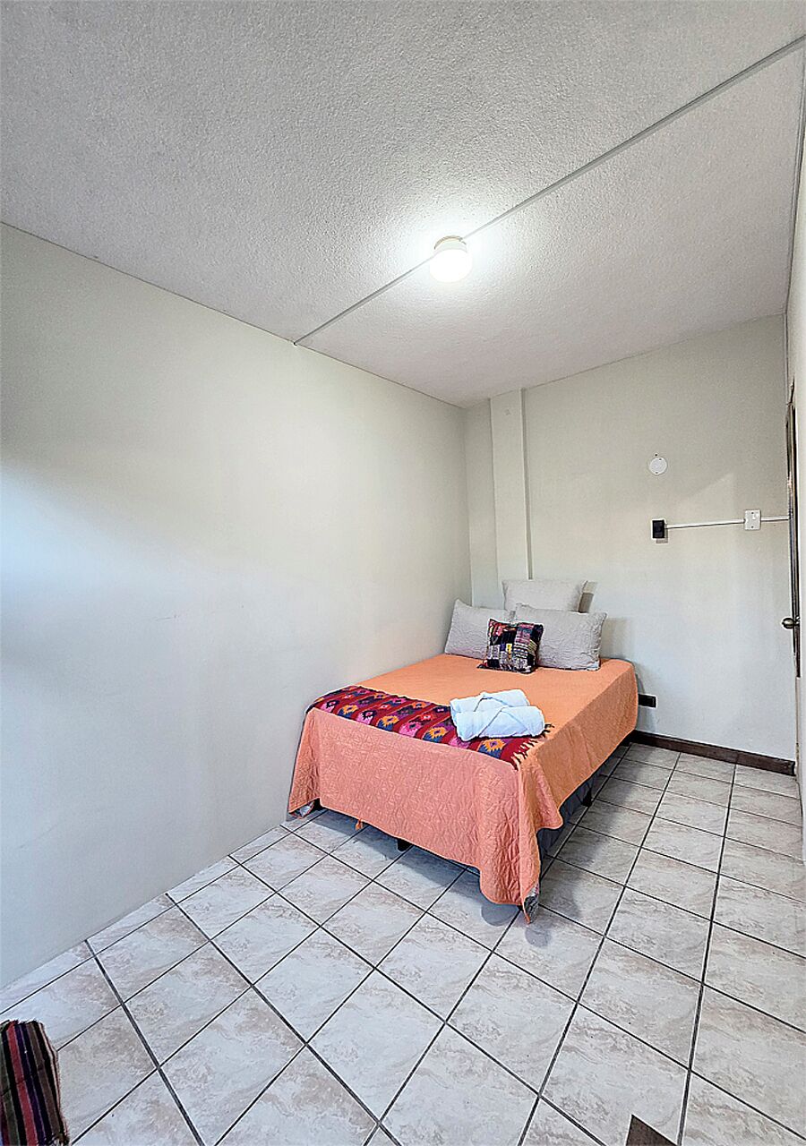 A small, single-bed room with a tiled floor, a plain white ceiling, and a simple bed with a pink blanket.