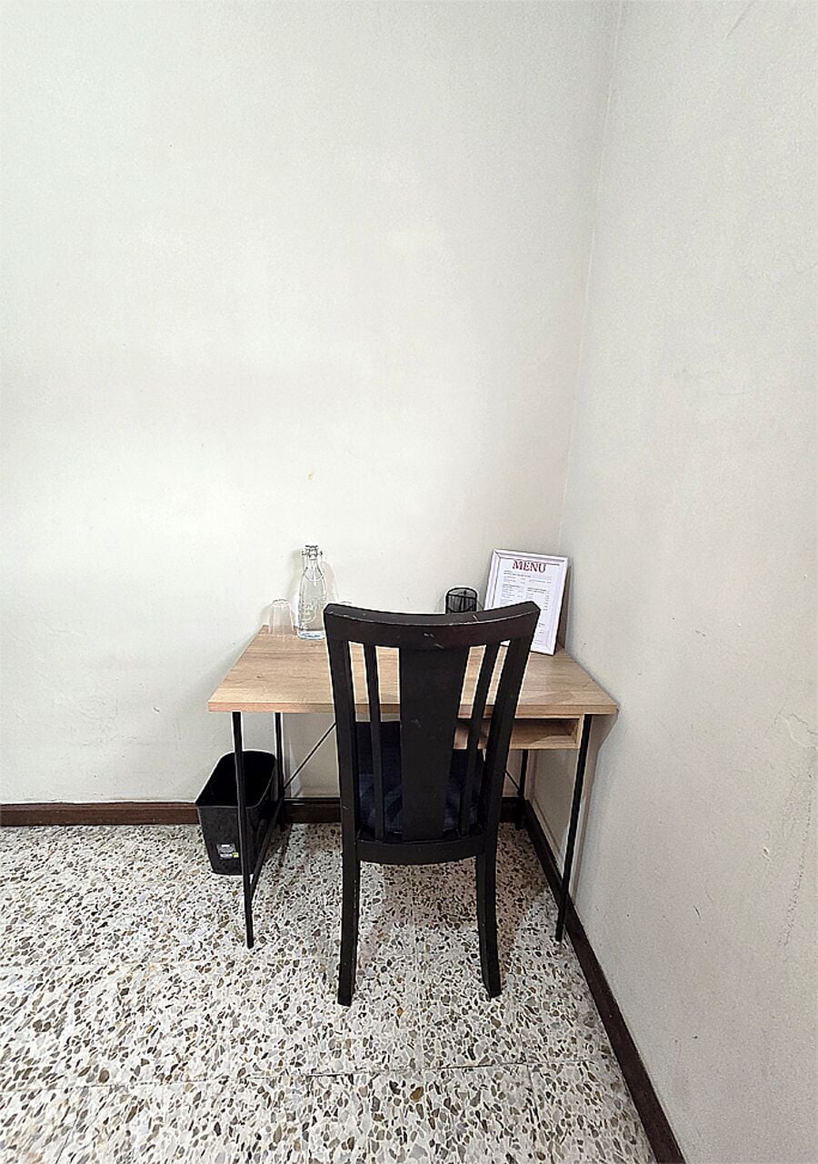 A corner room with a wooden table, a black chair, a glass bottle, and a menu sign.
