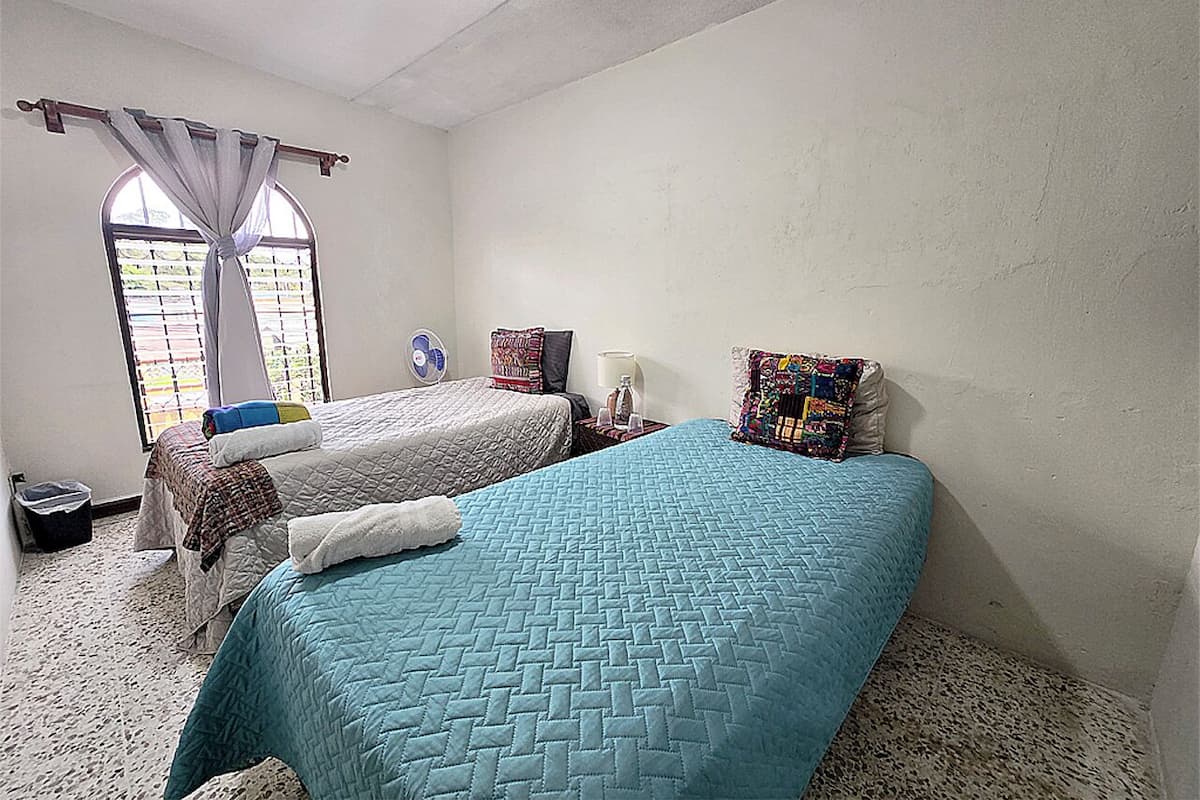 Une chambre avec deux lits, une fenêtre avec des rideaux, une petite table avec une lampe et une poubelle.