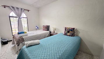 A bedroom with two beds, a window with curtains, a small table with a lamp, and a trash bin.