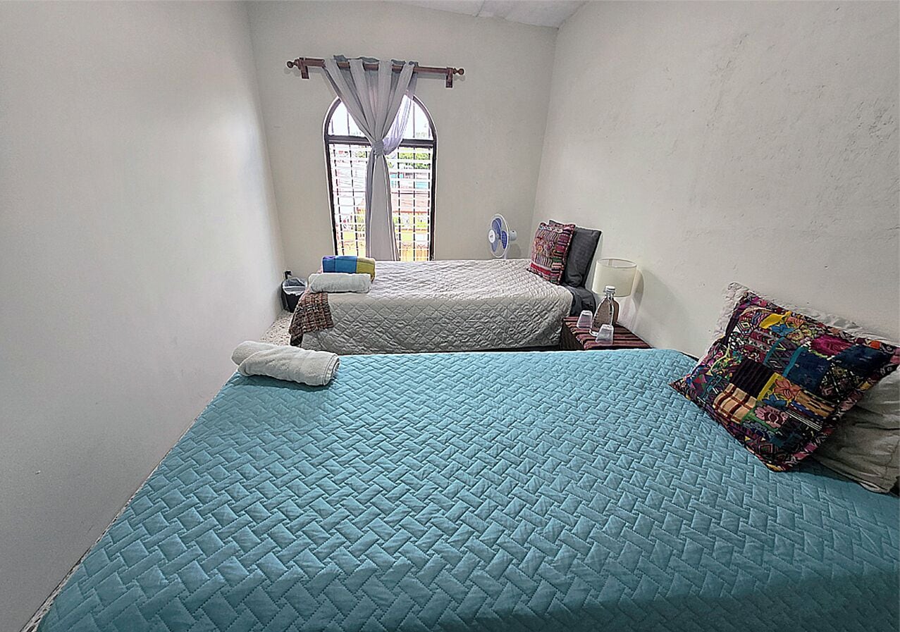 A bedroom with a bed, a window with curtains, a bedside table with a lamp, and a wall-mounted fan.