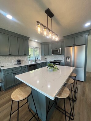 A modern kitchen with a large island, bar stools, a built-in microwave, and a stainless steel refrigerator. - CDA Ponderosa House (Coeur d'Alene)