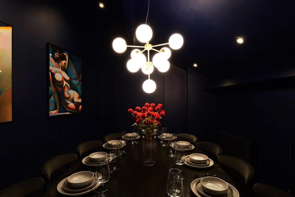 A dark dining room with a table set for six, a modern chandelier, and a painting on the wall.